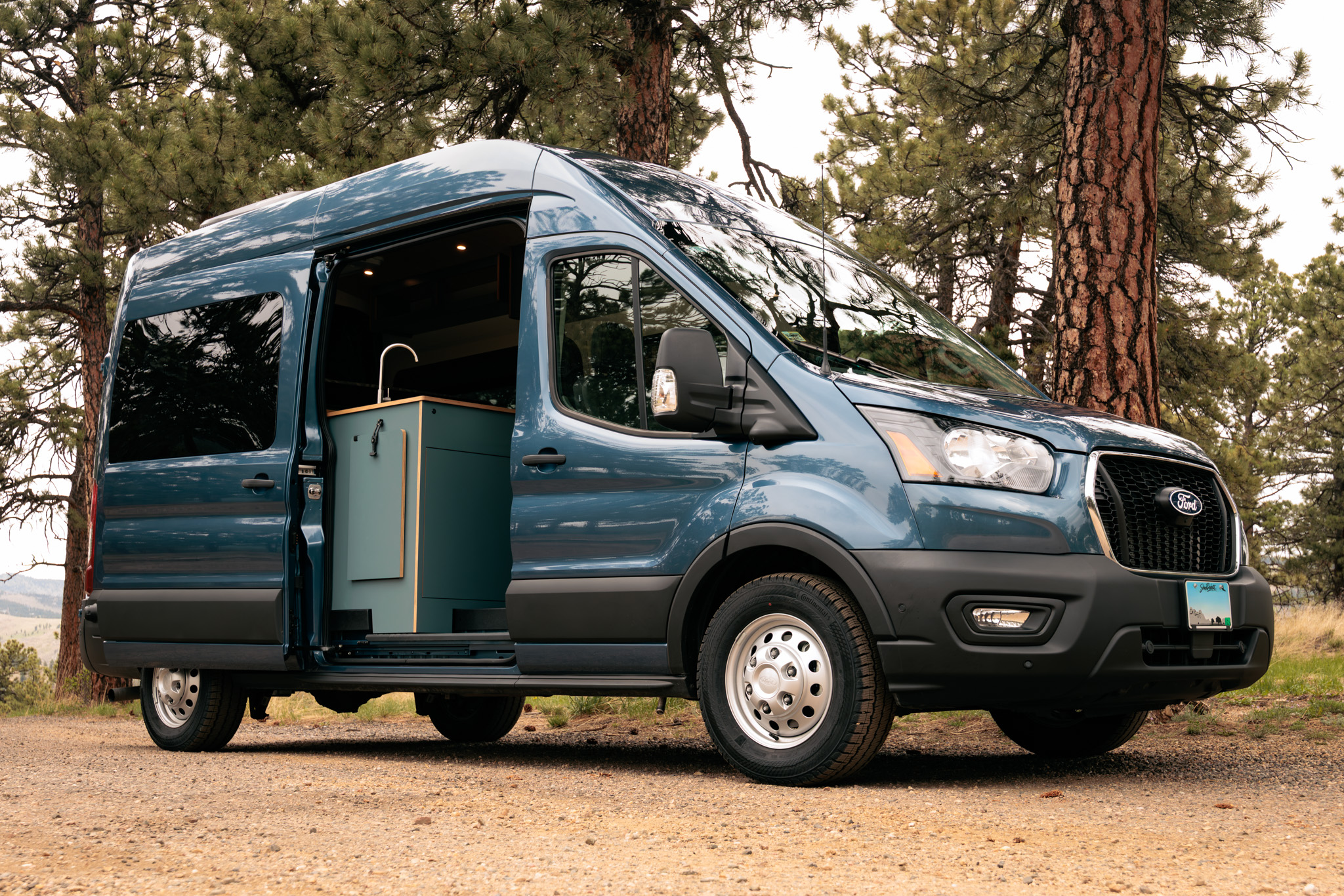 Blue Ford Transit Timber Rambler parked among pine trees, sliding door open on the galley
